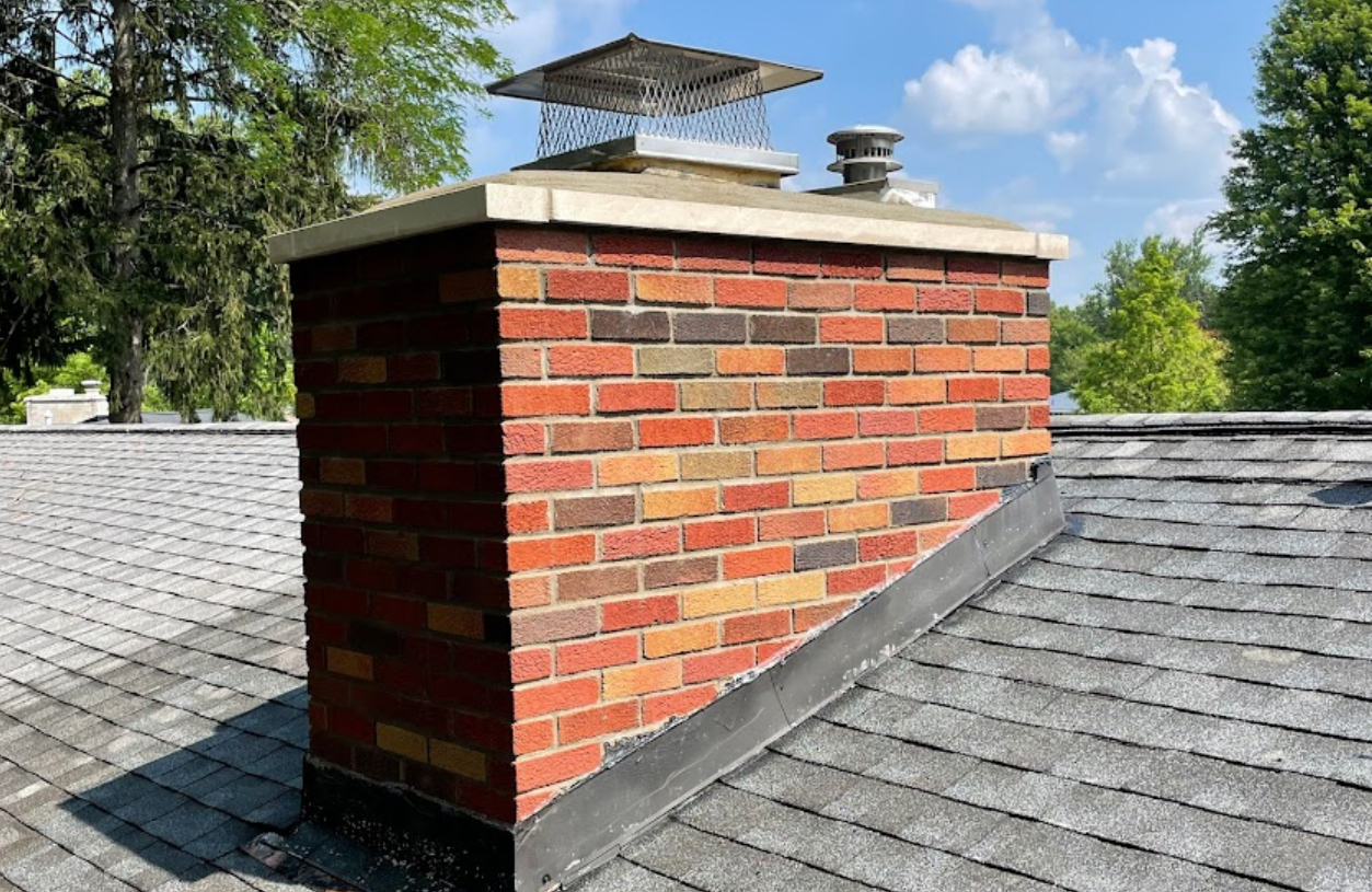 Rebuilt brick chimney with a concrete crown, metal cap and new step flashing at the asphalt shingle roof. Roofing work by Alba Pro Construction serving Wyckoff, NJ.