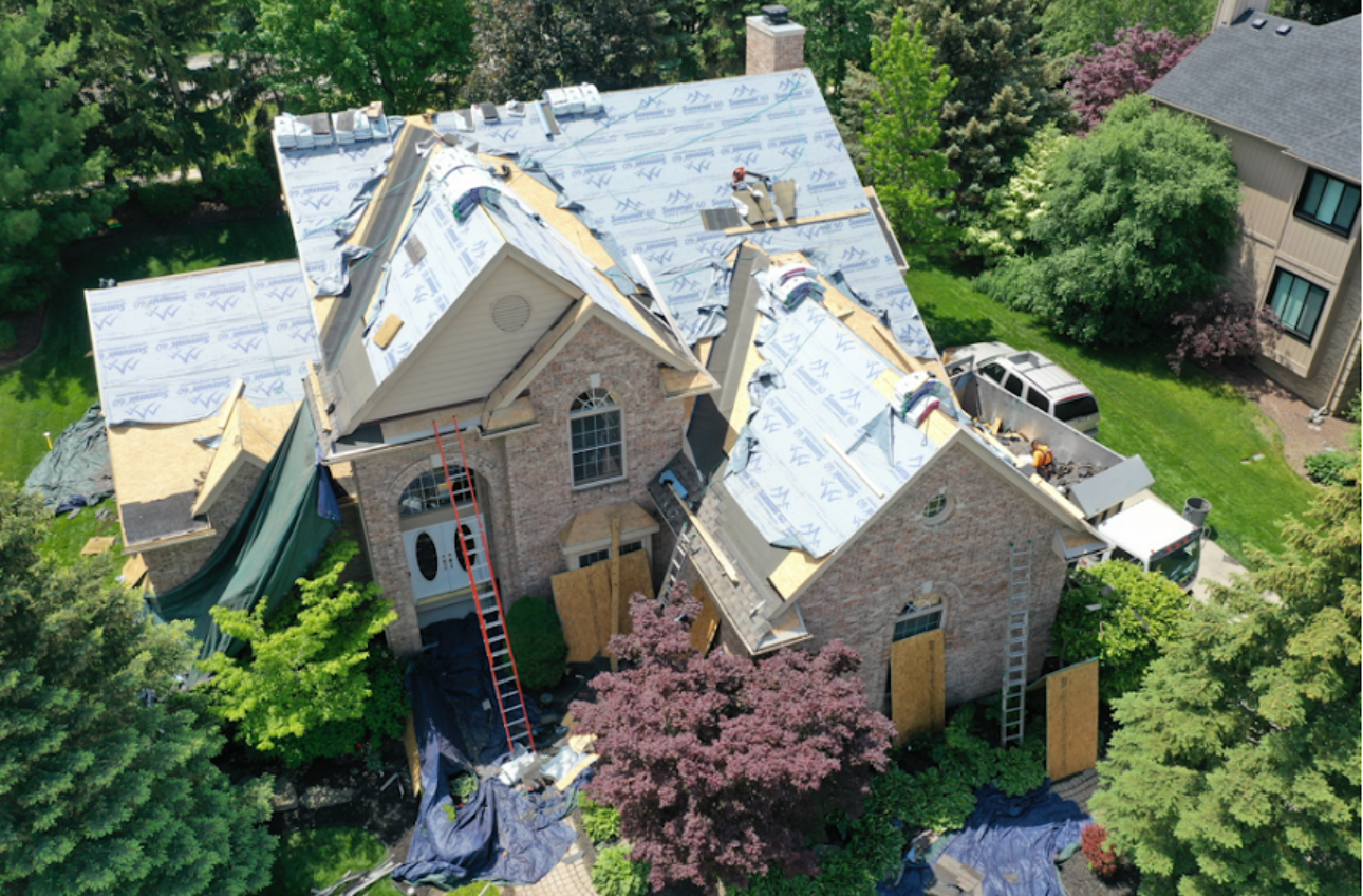 Aerial view of a large brick home with new decking and underlayment installed and tarps covering the shrubs below. Roofing work by Alba Pro Construction serving Englewood, NJ.