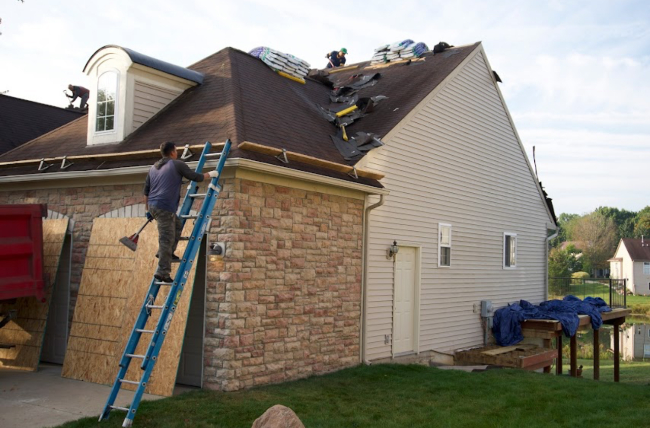 Roofer climbing a ladder while the crew strips underlayment from a stone and vinyl home. Roofing work by Alba Pro Construction serving Garfield, NJ.