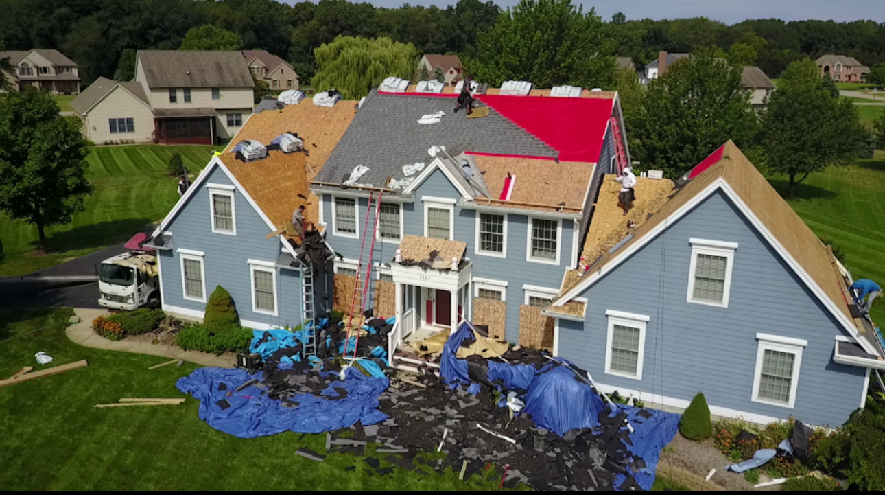 Aerial view of a blue colonial home mid roof replacement with new decking, red underlayment and tarps catching debris. Roofing work by Alba Pro Construction serving Oakland, NJ.