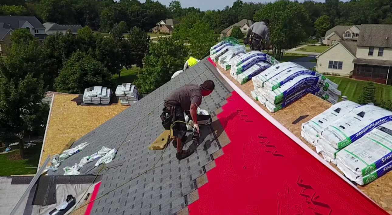 Drone view of a roofer nailing architectural shingles over red synthetic underlayment with shingle bundles staged along the ridge. Roofing work by Alba Pro Construction serving Newark, NJ.