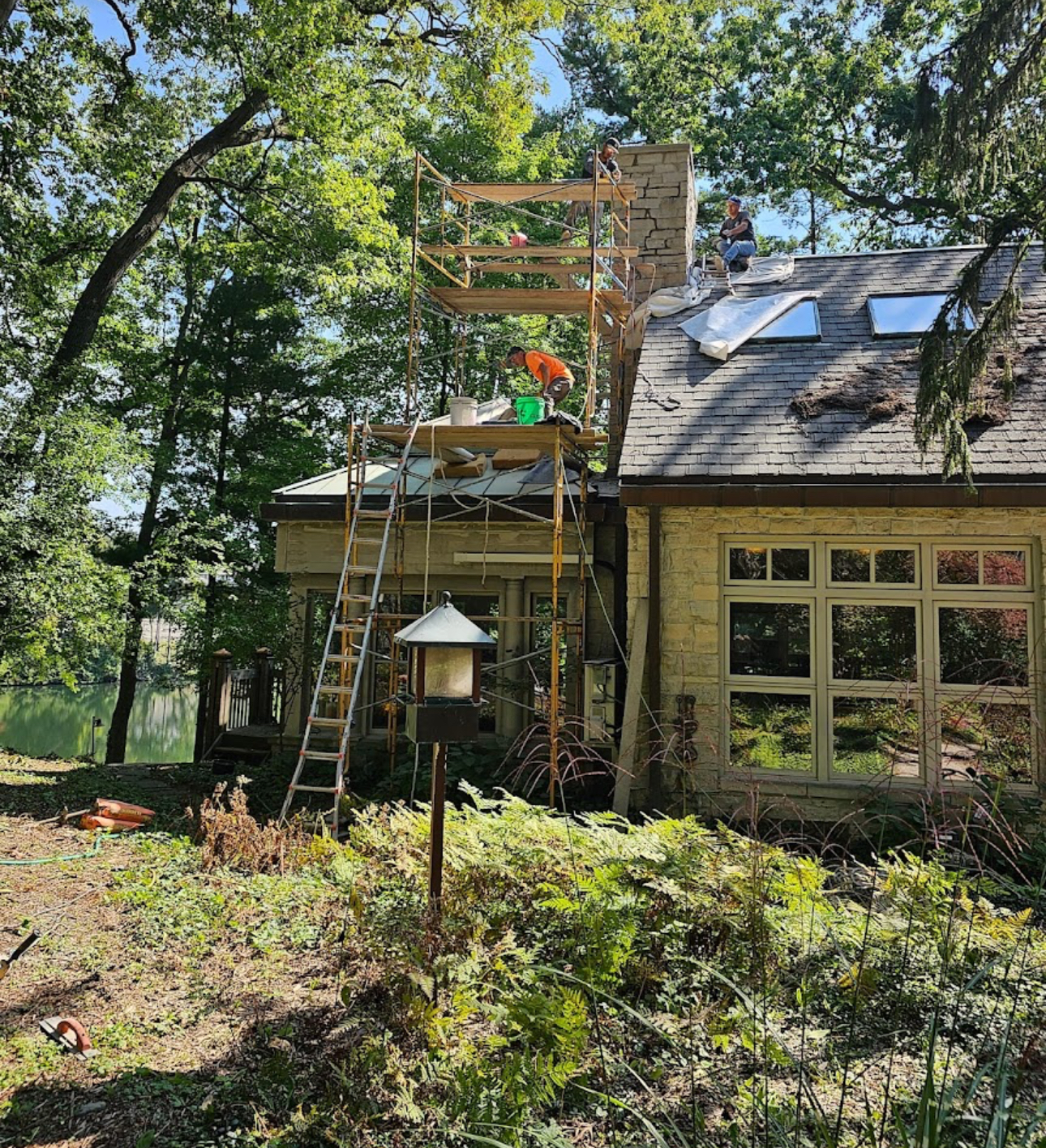 Crew working from scaffolding around a stone chimney during roof and chimney repairs on a wooded property. Roofing work by Alba Pro Construction serving Midland Park, NJ.
