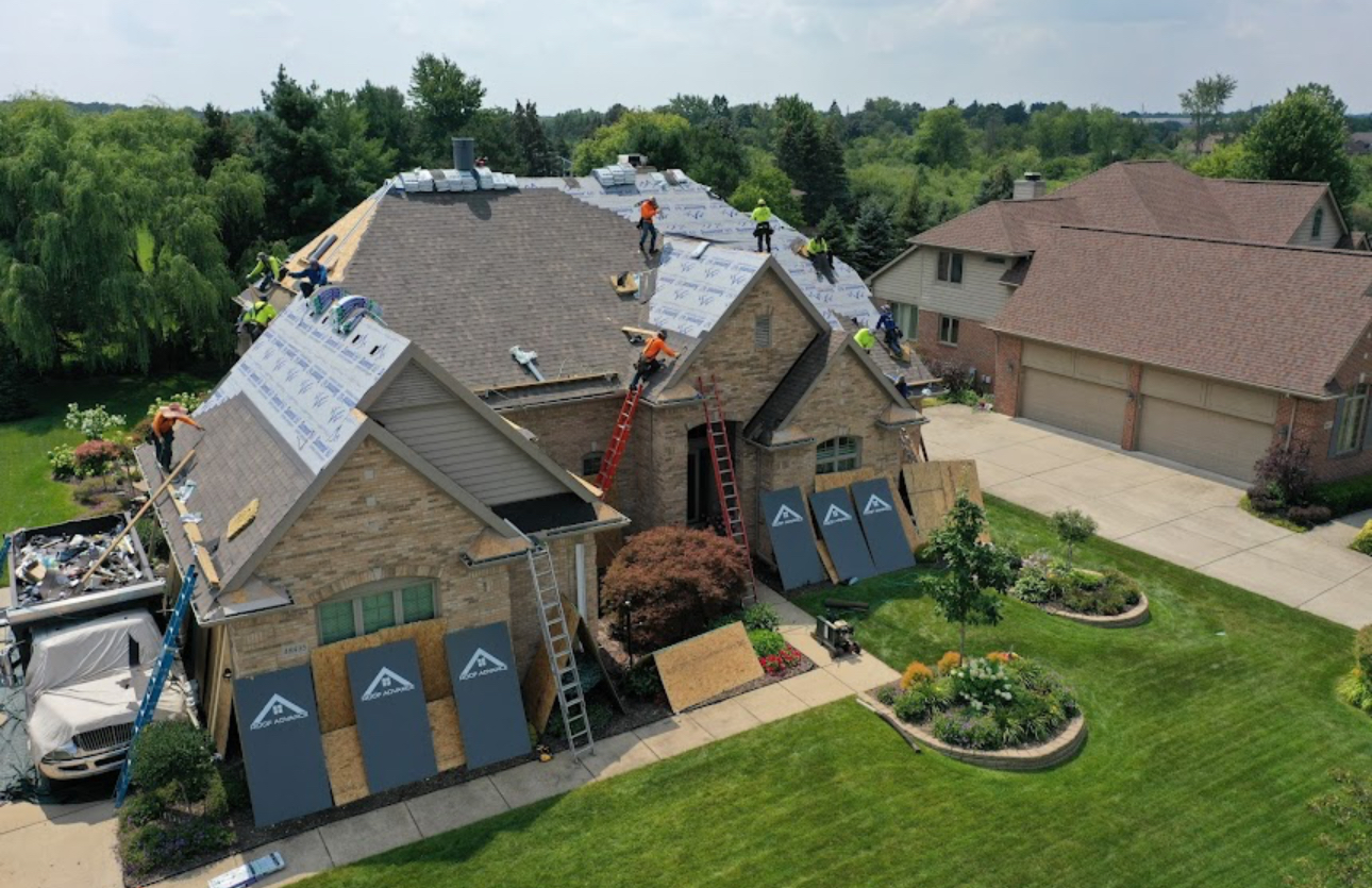 Aerial view of a roofing crew installing synthetic underlayment on a large brick home with plywood panels shielding the windows and landscaping. Roofing work by Alba Pro Construction serving Hackensack, NJ.